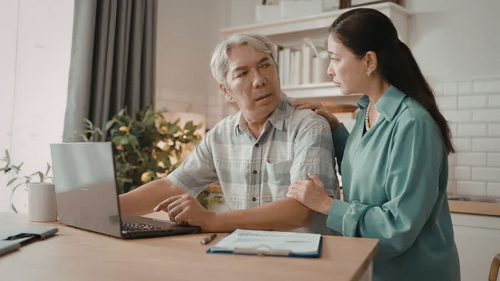Concerned woman comforts an older man sitting at a laptop with documents in their kitchen.